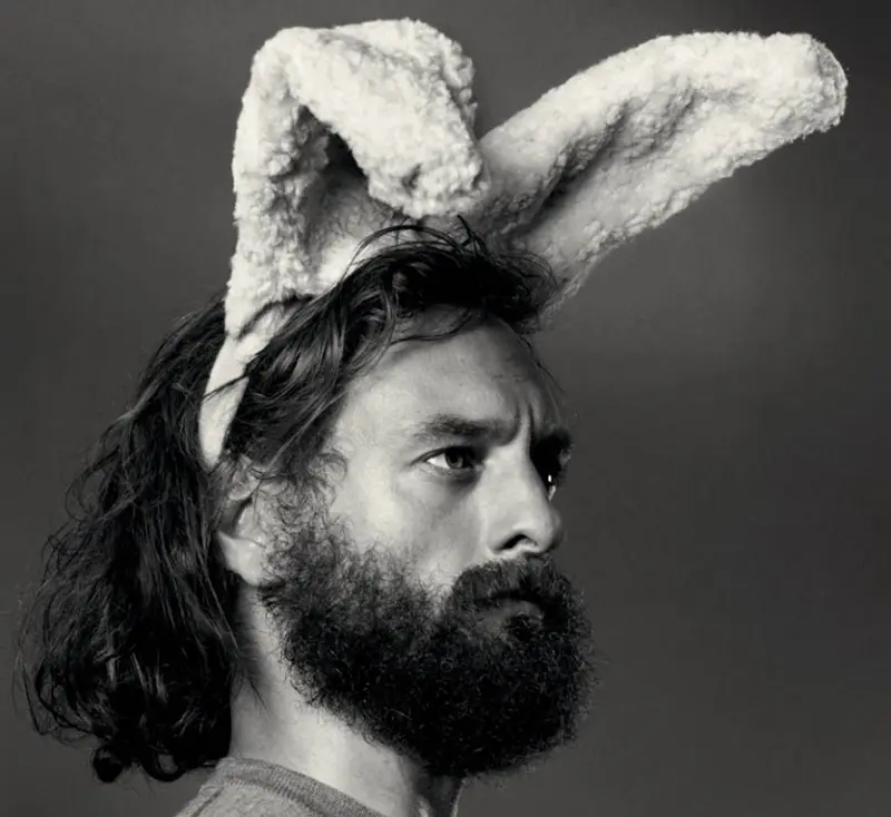Black-and-white portrait of a bearded man with long hair wearing fluffy bunny ears and a serious expression