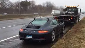 A black Porsche 911 broken down on a wet highway shoulder in front of a tow truck