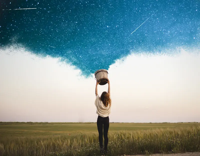 Woman in a wheat field holding a bucket overhead as a starry night sky pours out of it