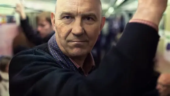 A tired, grim-faced middle-aged man holding the handrail in a crowded subway carriage