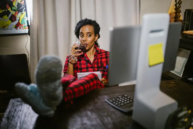 Woman in red flannel pyjamas sipping red wine at her desk, feet up, notepad in hand