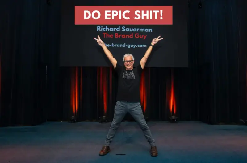 Richard Sauerman on stage, arms raised in peace signs, under a 'Do Epic Shit!' sign