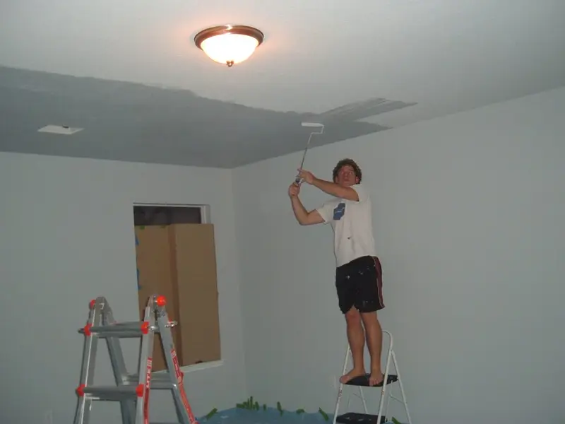 A young man on a step stool painting a ceiling with a roller
